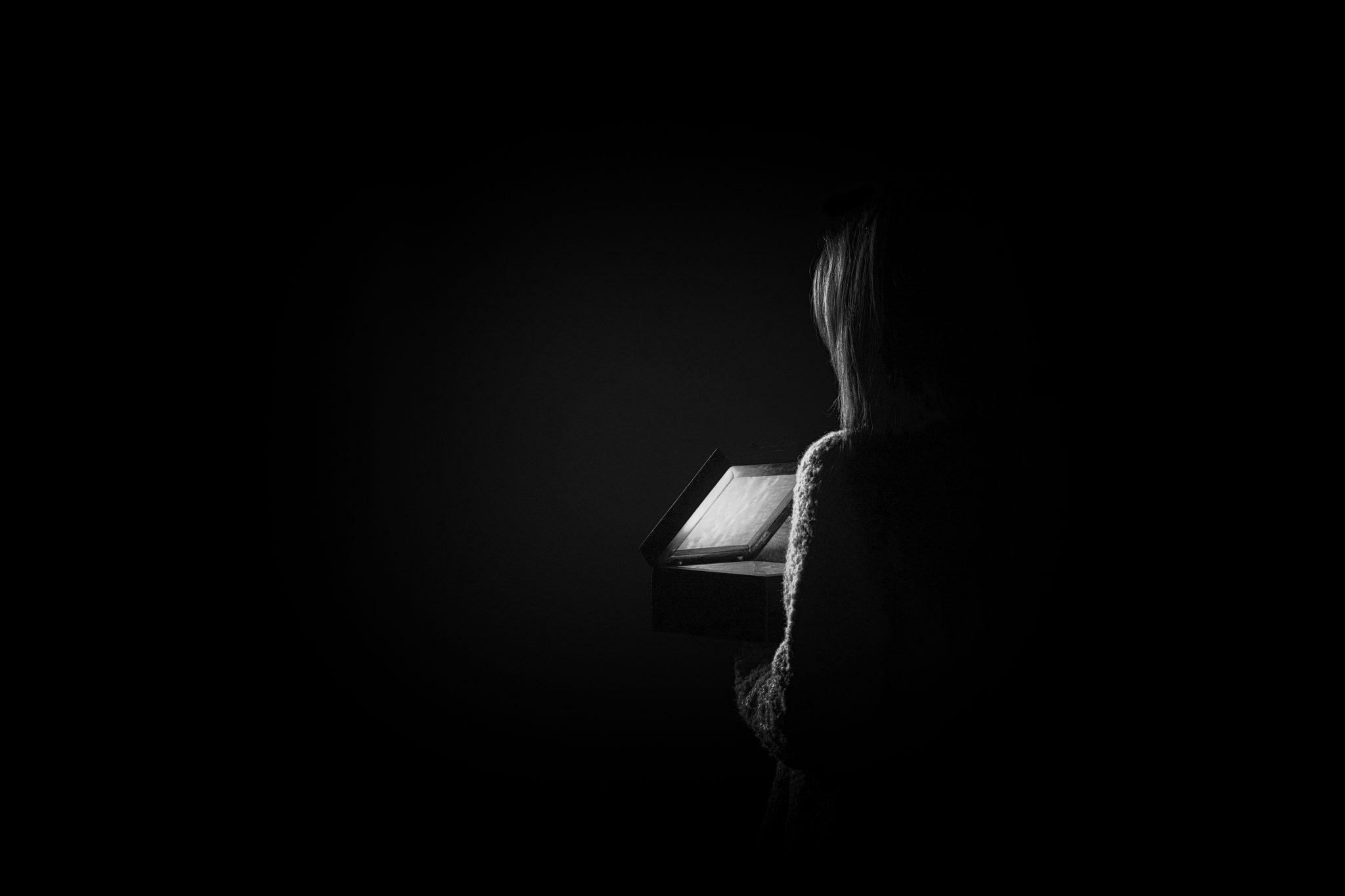Black and white photo of a woman, from behind, looking into a box with a glowing light inside.