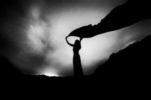 Black and white photo of a woman's sillouette holding a long scarf high over her head against a dawning sky.
