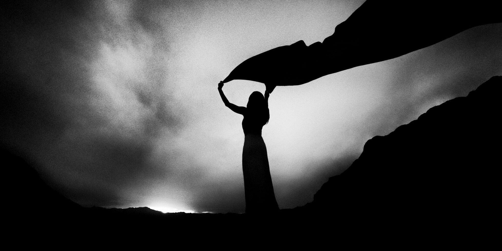 Black and white photo of a woman's sillouette holding a long scarf high over her head against a dawning sky.